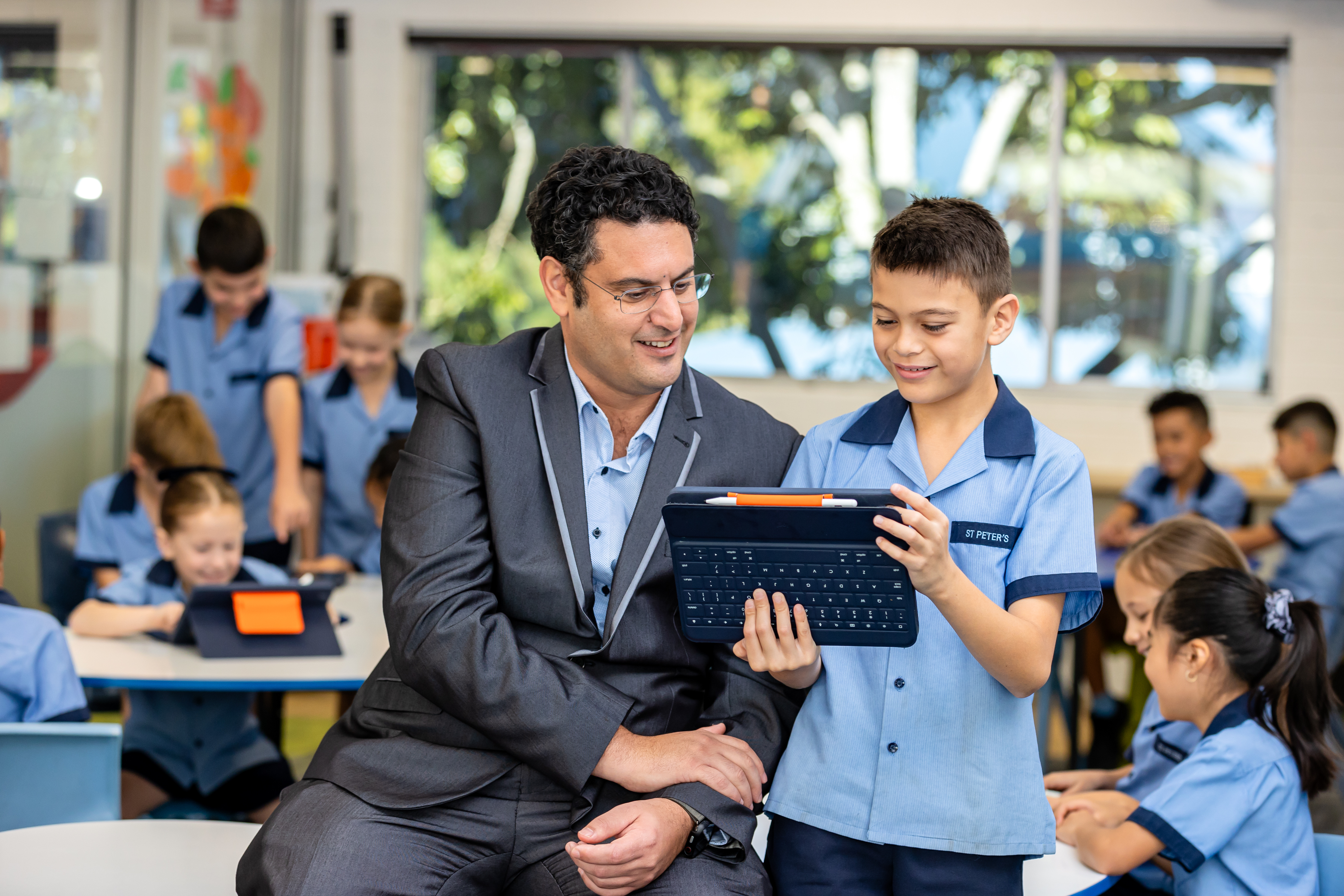 Teacher supporting a student using a tablet during a classroom learning activity with other students engaged in digital learning in the background Provide your feedback on BizChat