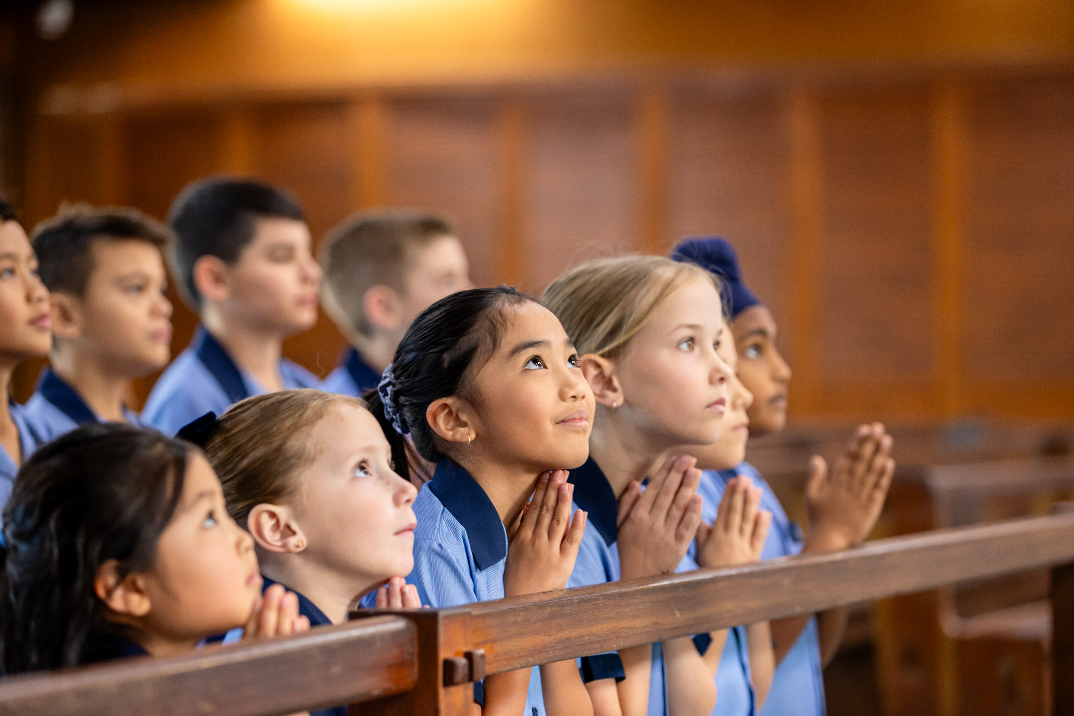 Students in school uniform kneeling in a church pew with hands together in prayer during a Catholic liturgy Provide your feedback on BizChat