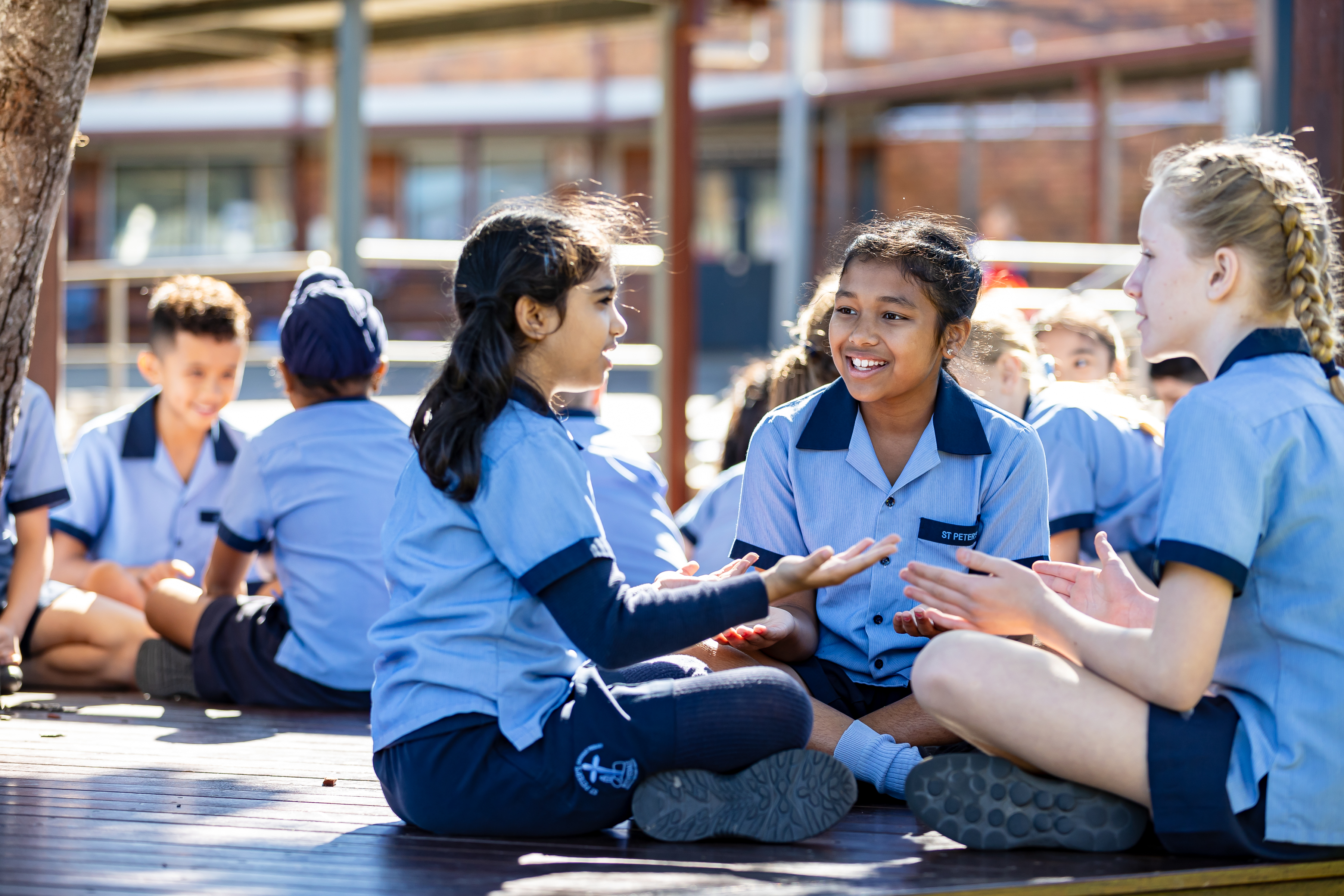 Students in school uniform sitting together outdoors on a deck, playing a hand game and interacting as part of social play during break time