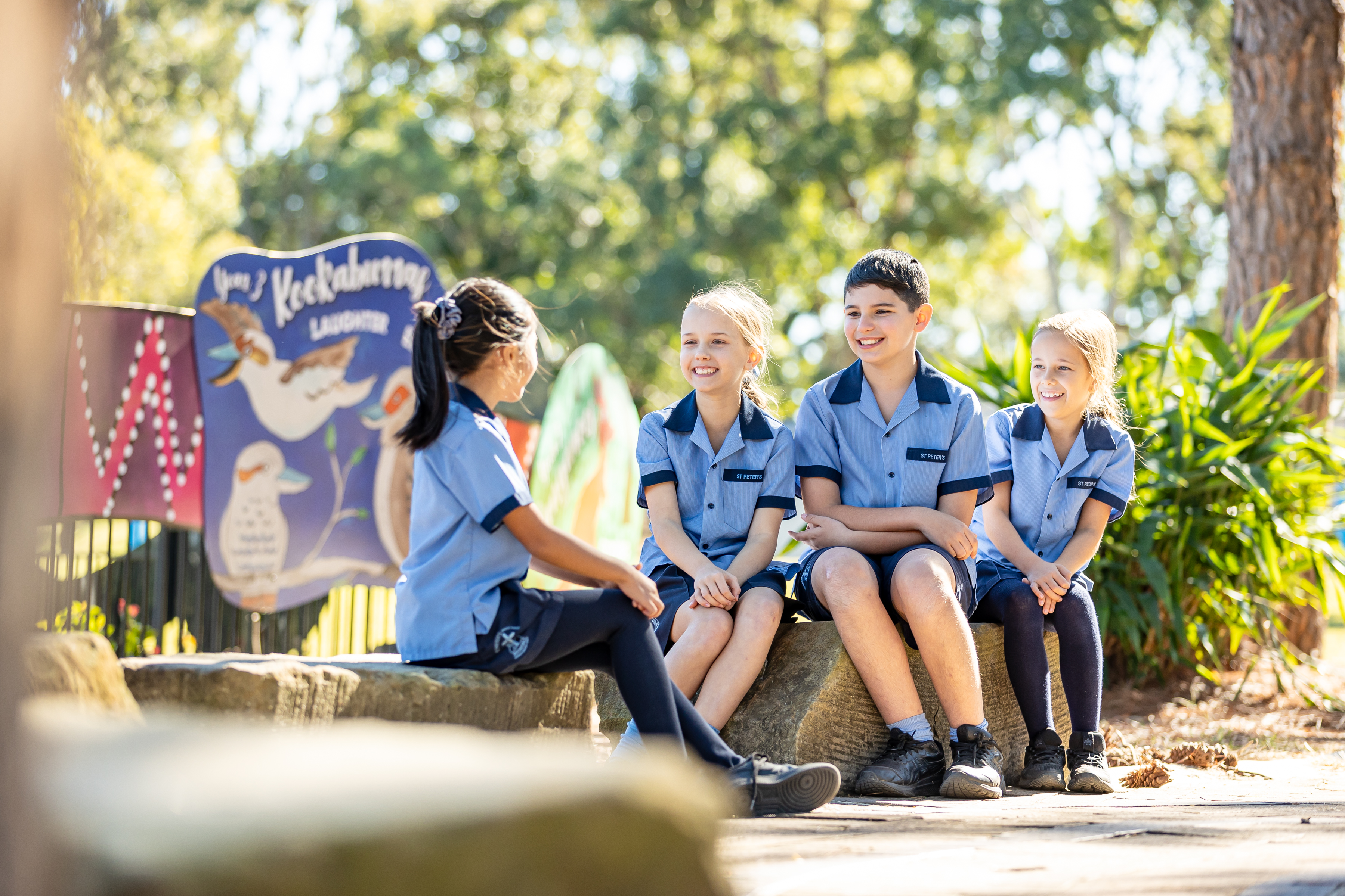 Students in school uniform sitting together outdoors on rocks near a playground feature, engaging in conversation and social interaction during break time